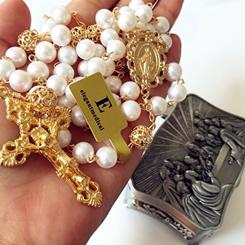 Hand holding a golden rosary with pearl beads near a decorative box.