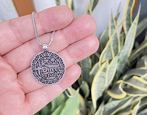 Hand holding a circular pendant with engraved text, plant in background.