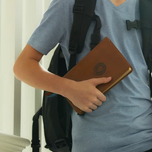 Person holding a brown notebook and wearing a backpack
