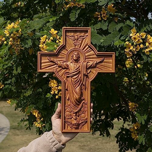 Hand holding a wooden cross with Jesus, against a background of green leaves and yellow flowers