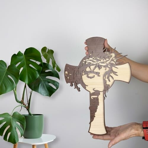 Hand holding a wooden cross with Jesus design next to a potted plant.
