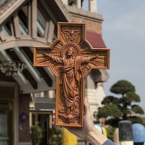 A hand holding a wooden crucifix outdoors with a building in the background